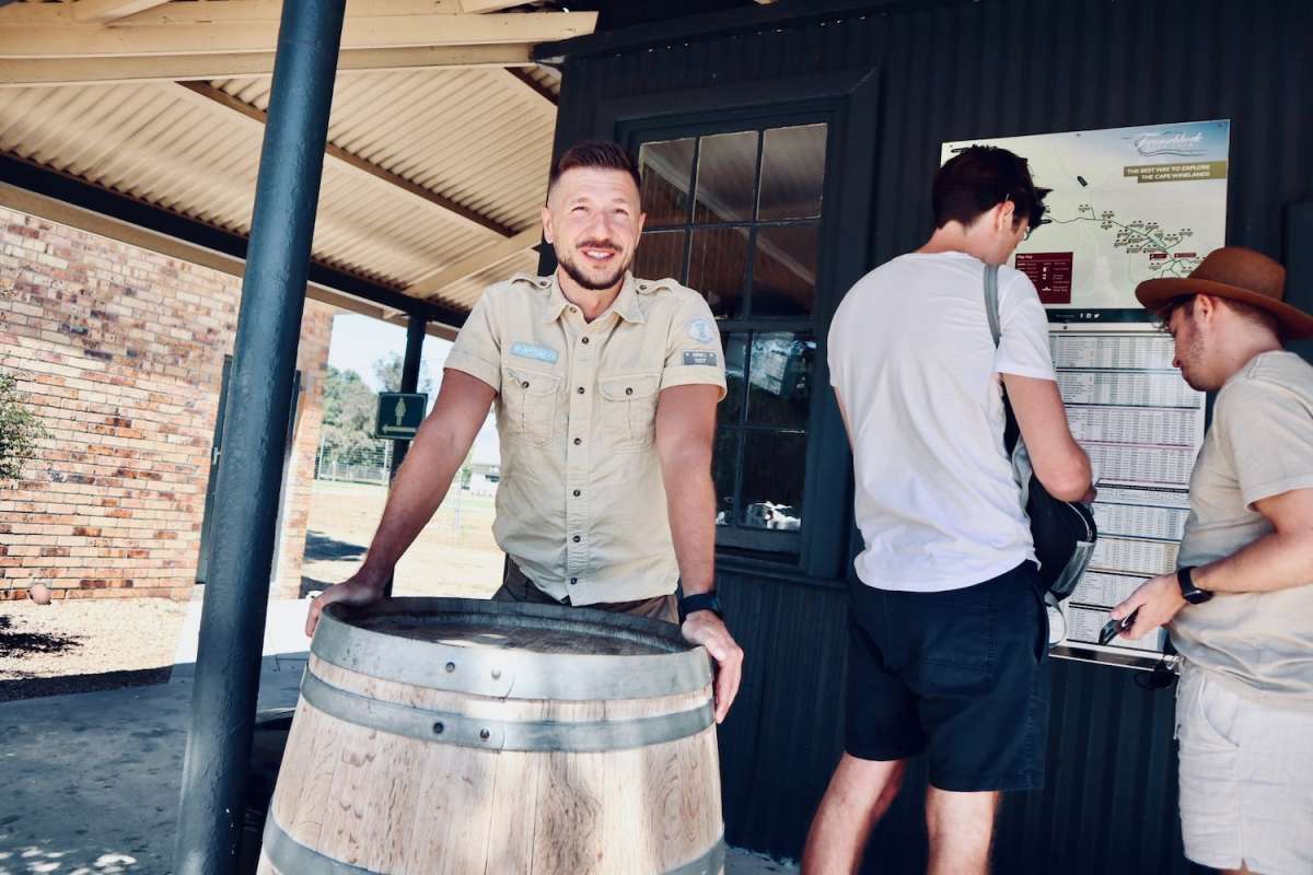 A cheerful man leaning on a wooden barrel at a winery in Franschhoek near gay Cape Town, with a wine tram timetable in the background, showcasing the region's wine culture.