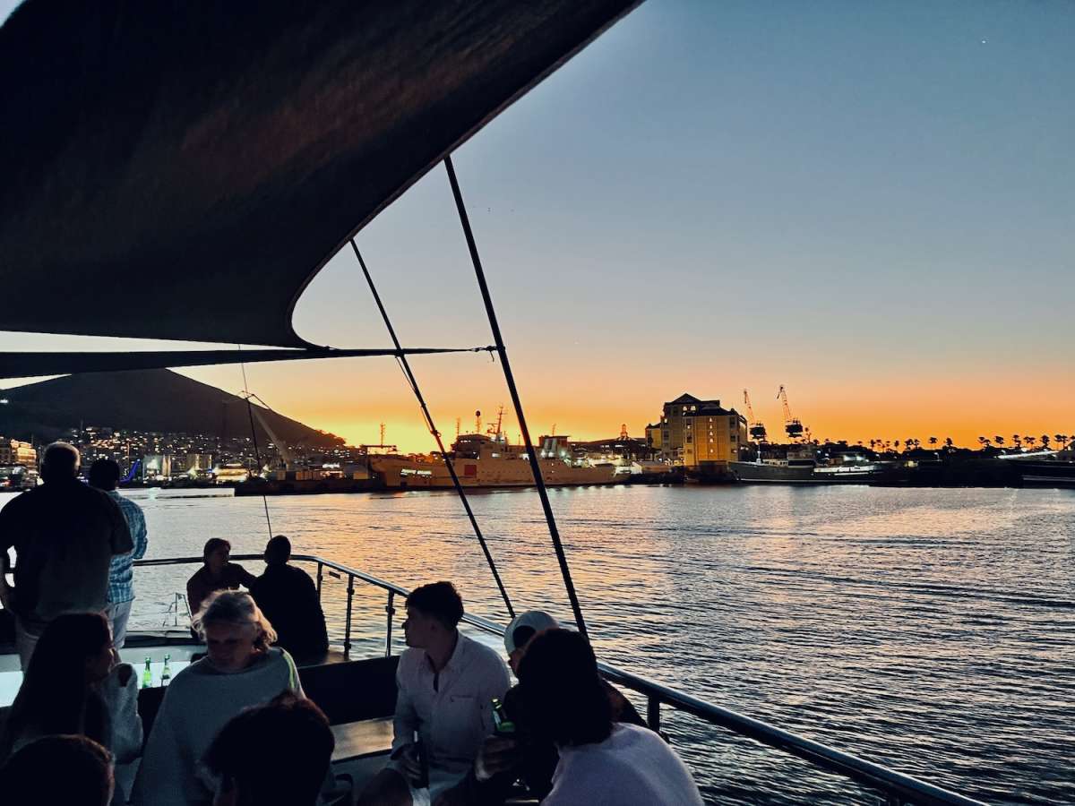 Sunset cruise view at the V&A Waterfront in gay Cape Town, with silhouettes of people against a backdrop of illuminated ships and palm trees, highlighting the city's nightlife.