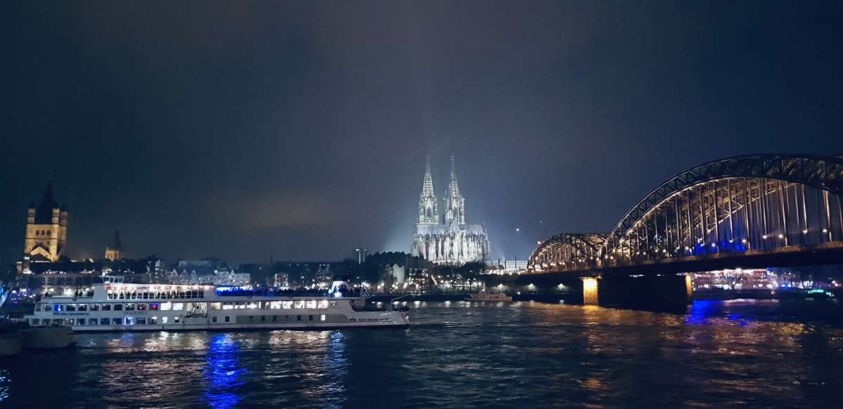 Cologne's iconic Gothic Cathedral stands illuminated against the night sky, with the Hohenzollern Bridge aglow and a river cruise boat sailing the Rhine, reflecting the vibrant nightlife of gay Cologne.