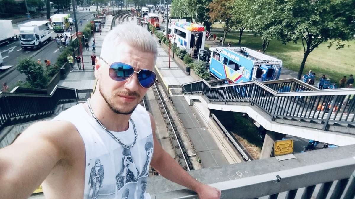 A man in a sleeveless shirt and aviator sunglasses takes a selfie with Cologne's bustling street below, showcasing the energetic atmosphere of gay Cologne during a city event.