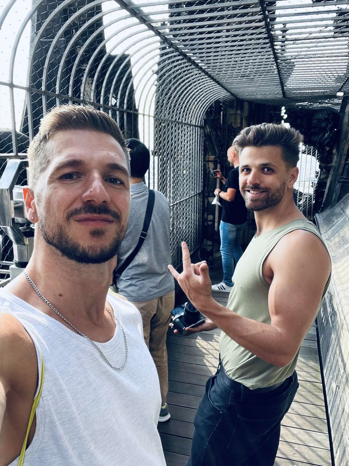 Two men in casual summer wear share a playful moment in a cage-like tunnel, capturing the camaraderie and lively spirit prevalent in the gay Cologne community.