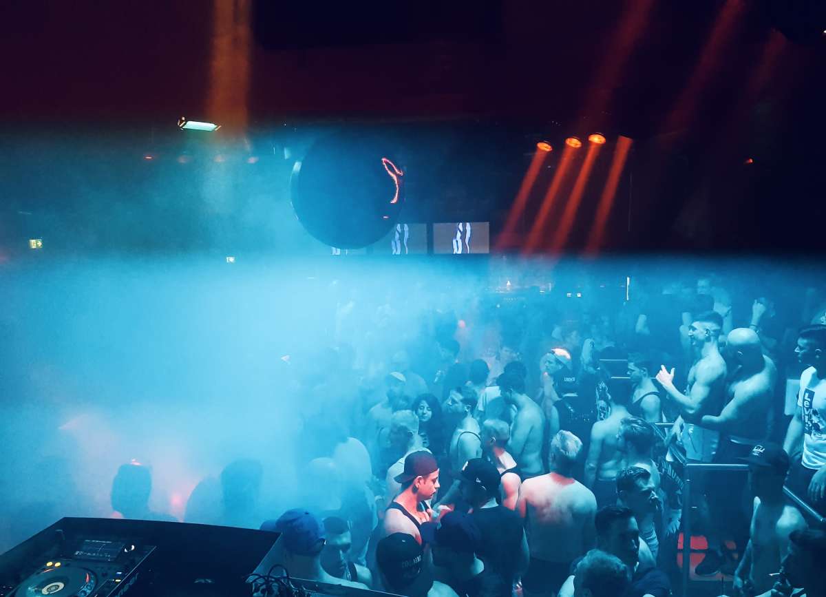 A packed dance floor with blue lighting at Green Komm, one of Cologne's famous gay parties, where attendees are immersed in a sea of fog and revelry.