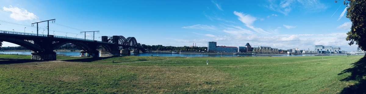 Panoramic view of the Rhine River in Cologne, with the South Bridge foregrounding the serene waters and urban skyline, a peaceful yet vibrant part of gay Cologne.