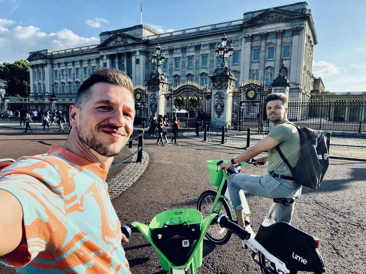 Two men enjoying a bike ride near Buckingham Palace, with one man playfully sticking out his tongue, showcasing Gay London's fun and lively cultural scene.