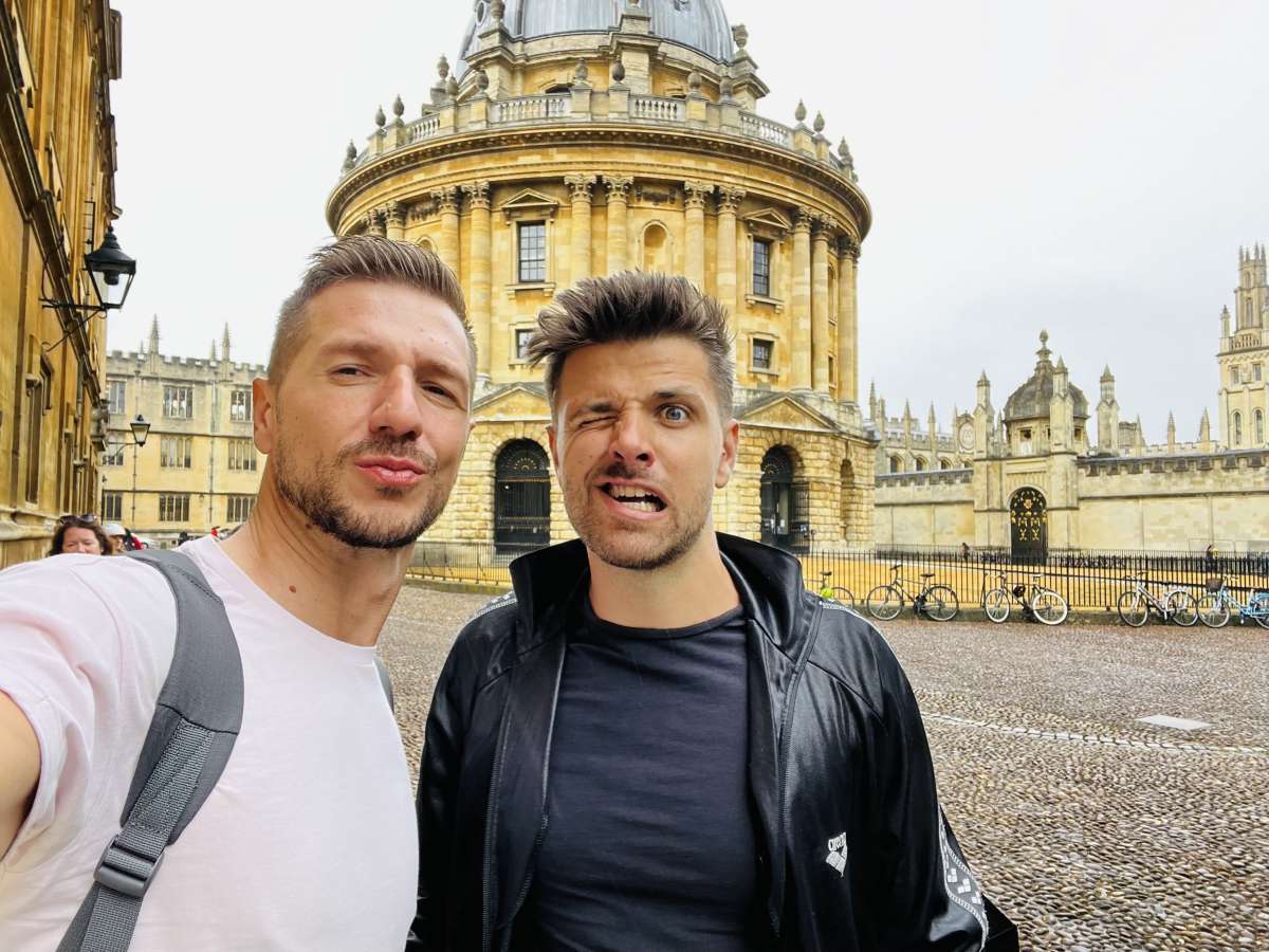 Two men making faces for a selfie with the grandiose Oxford University buildings in the background, a humorous take on Gay London's academic excursions.