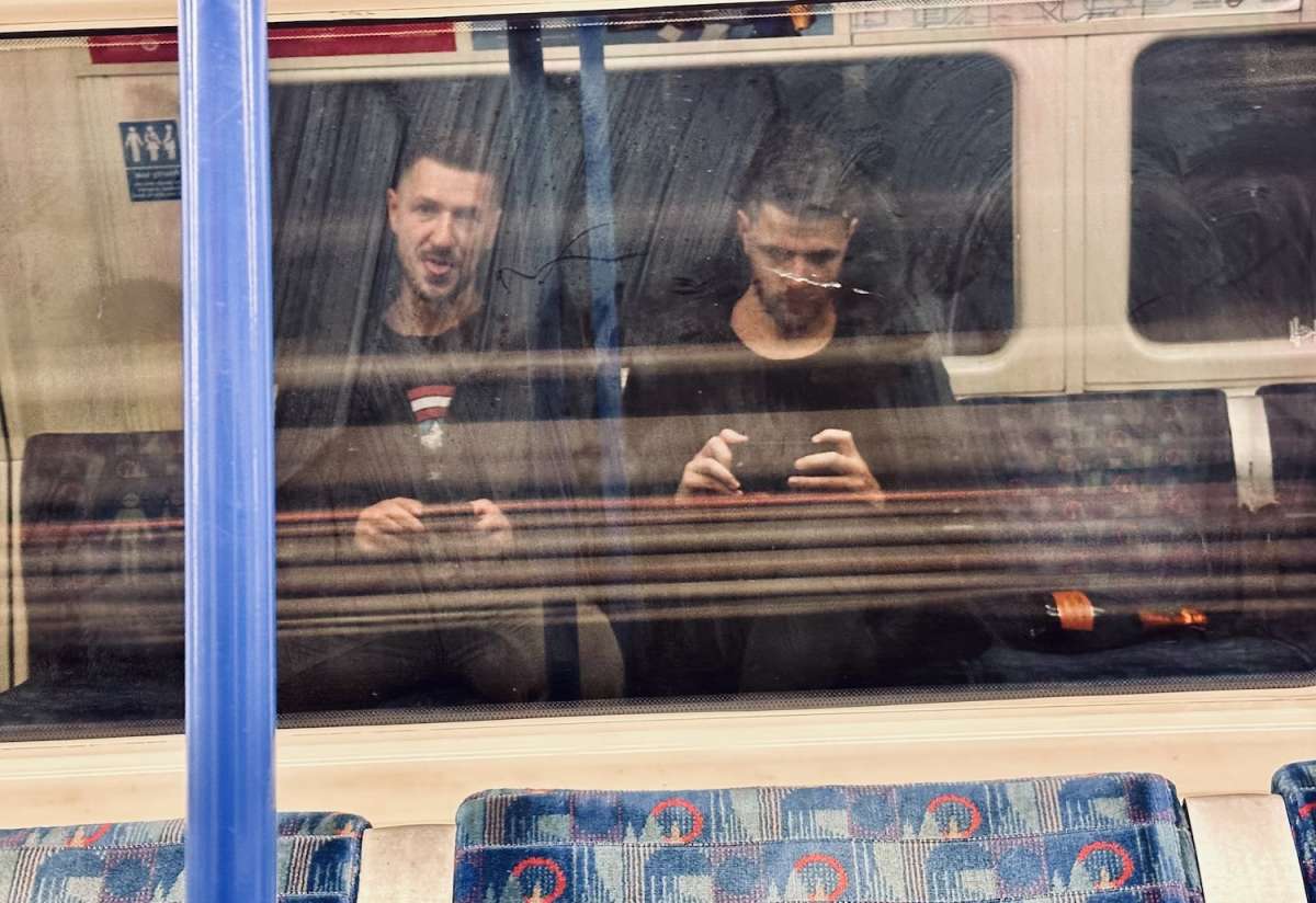 Reflection of two men sitting opposite each other in a London Tube carriage, a candid snapshot of everyday life in Gay London.