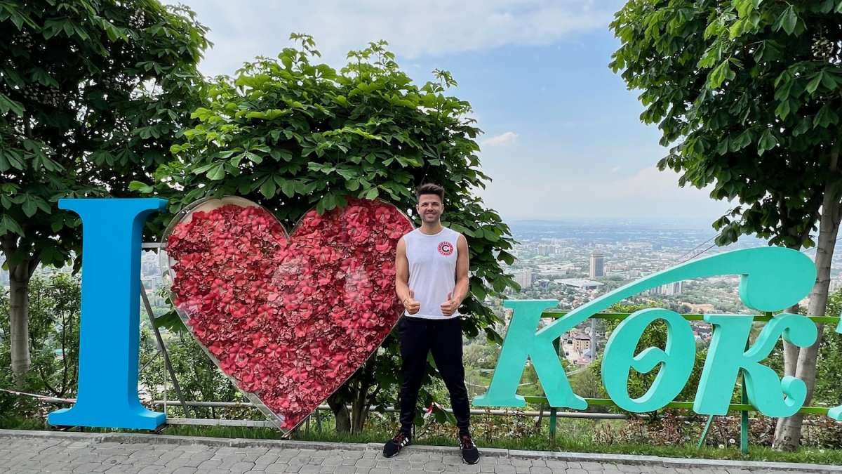 A cheerful man standing in front of the iconic 'I Love Kok Tobe' sign in Almaty, Kazakhstan, with a scenic city view in the background.