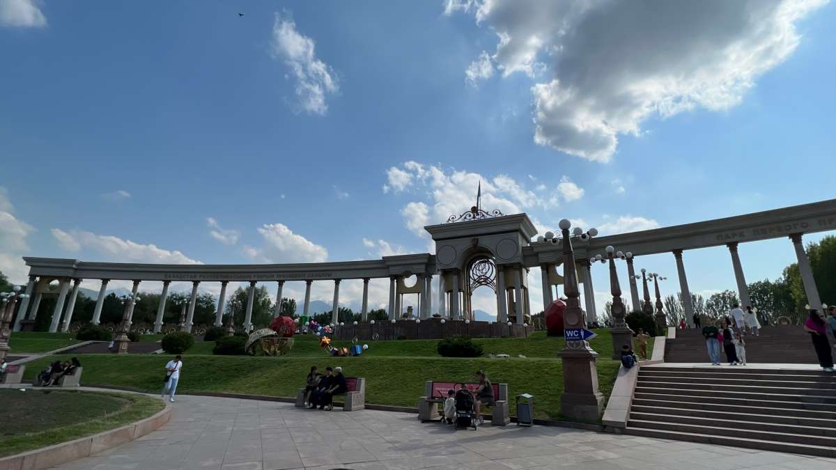 First President Park in Almaty, Kazakhstan, featuring a grand colonnade and green lawns, with people enjoying a sunny day outdoors.