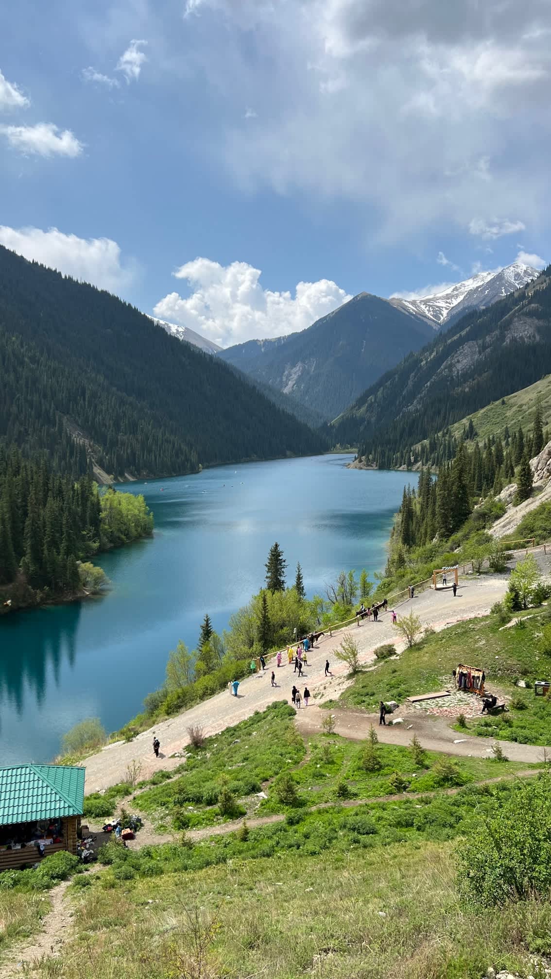 Serene view of Kolsay Lake, nestled among pine-covered hills in Kazakhstan, with visitors enjoying the pristine natural setting near Almaty.