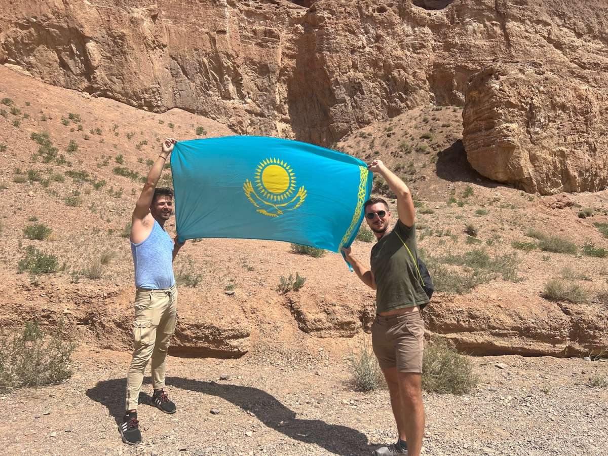 Two joyful men holding up the national flag of Kazakhstan in front of a rugged desert landscape, symbolizing pride and adventure near Almaty.