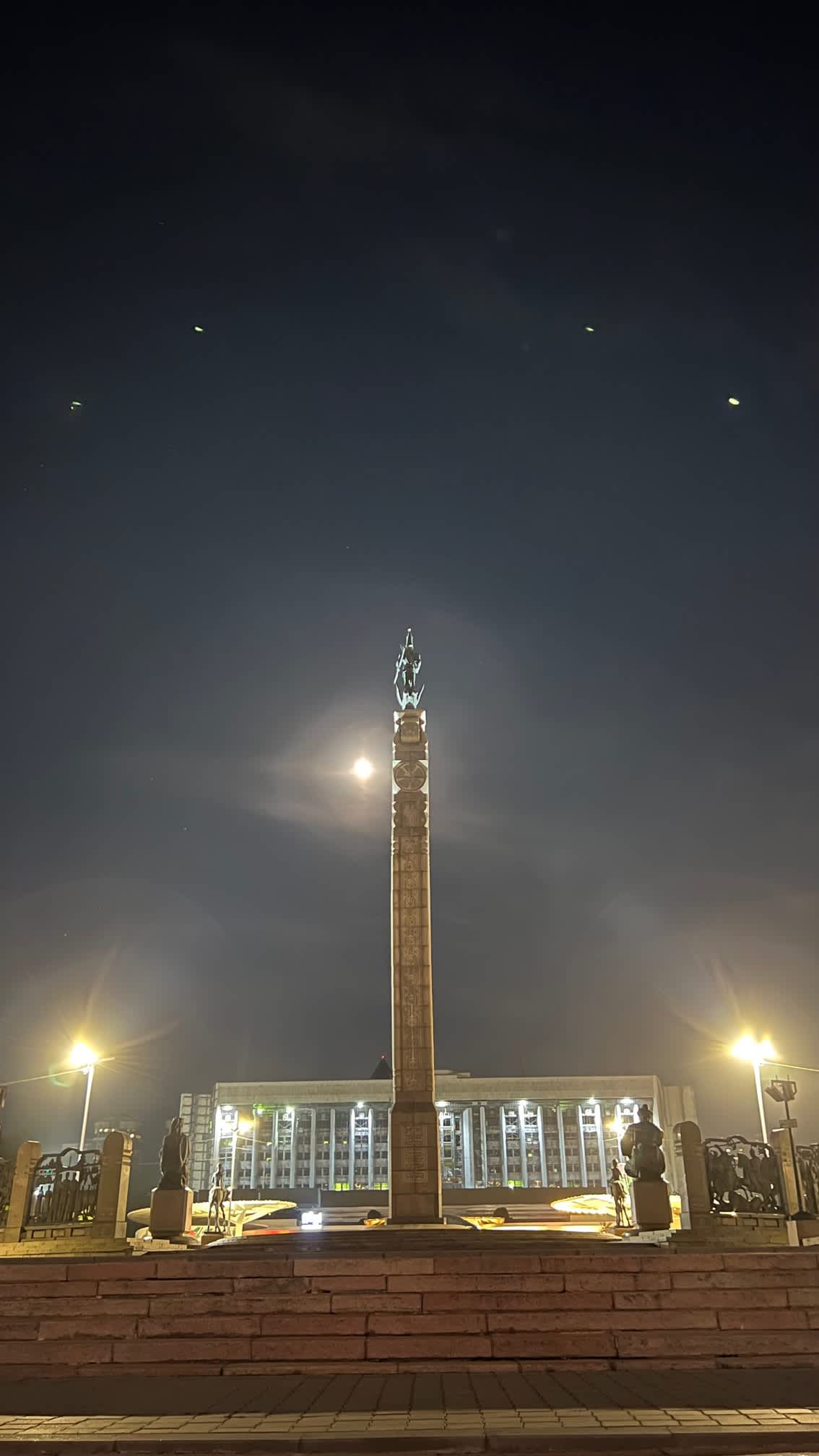 The Independence Monument in Almaty, Kazakhstan, illuminated at night with a full moon overhead, creating a dramatic urban scene.