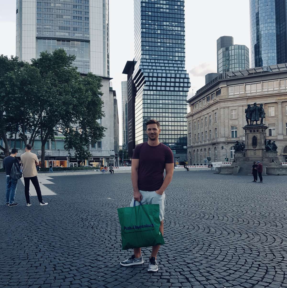 Smiling man holding a shopping bag stands in gay Frankfurt's central square, with the iconic Deutsche Bank towers in the background.