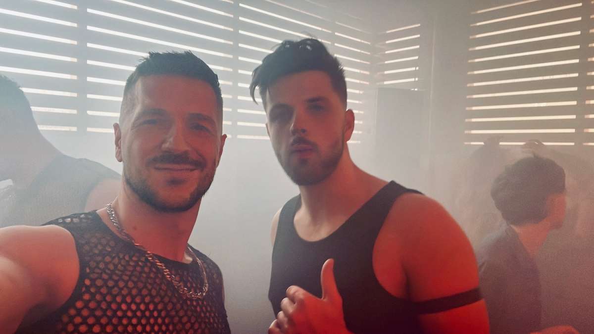 Two happy men taking a selfie with a smoky, backlit ambiance at Adonis, renowned as one of the best gay clubs in London.