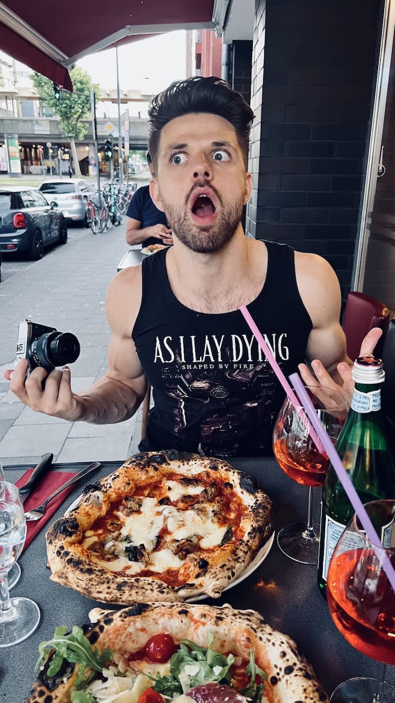 A surprised diner with an 'As I Lay Dying' tank top reacting to the sight of two artisanal pizzas at Toto & Pepino, a contender for the best restaurant in Cologne, set against an urban street backdrop.