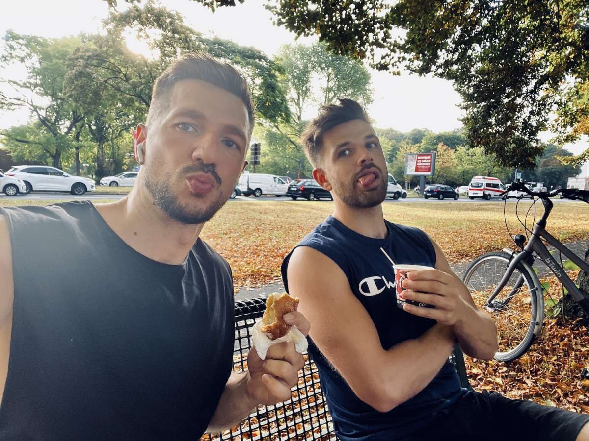 Two men enjoying a casual outdoor meal in a park in Cologne, with one savoring a croissant and the other drinking from a cup, representing the laid-back dining culture around some of the best restaurants in Cologne.