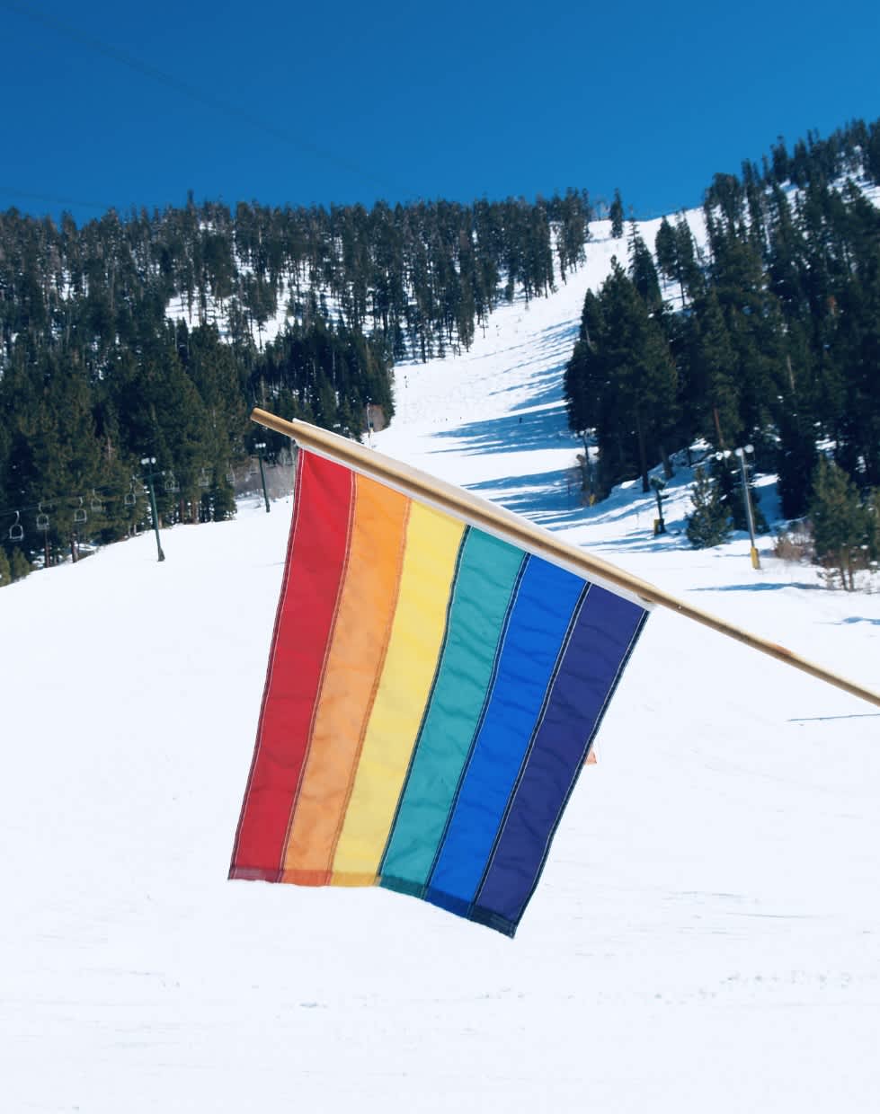 A vibrant LGBTQ+ rainbow flag prominently displayed against a snowy mountain slope, under a clear blue sky, symbolizing inclusion during Gay Ski Week.