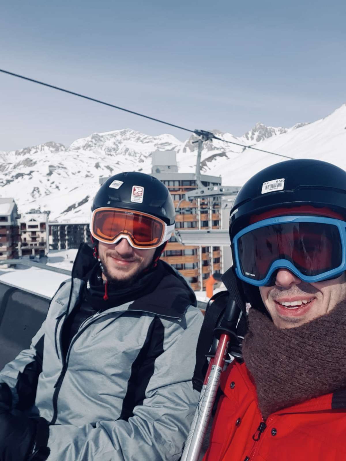 Two joyful skiers wearing goggles and winter gear, sharing a selfie moment with the snowy Tignes resort in the background, during Gay Ski Week.