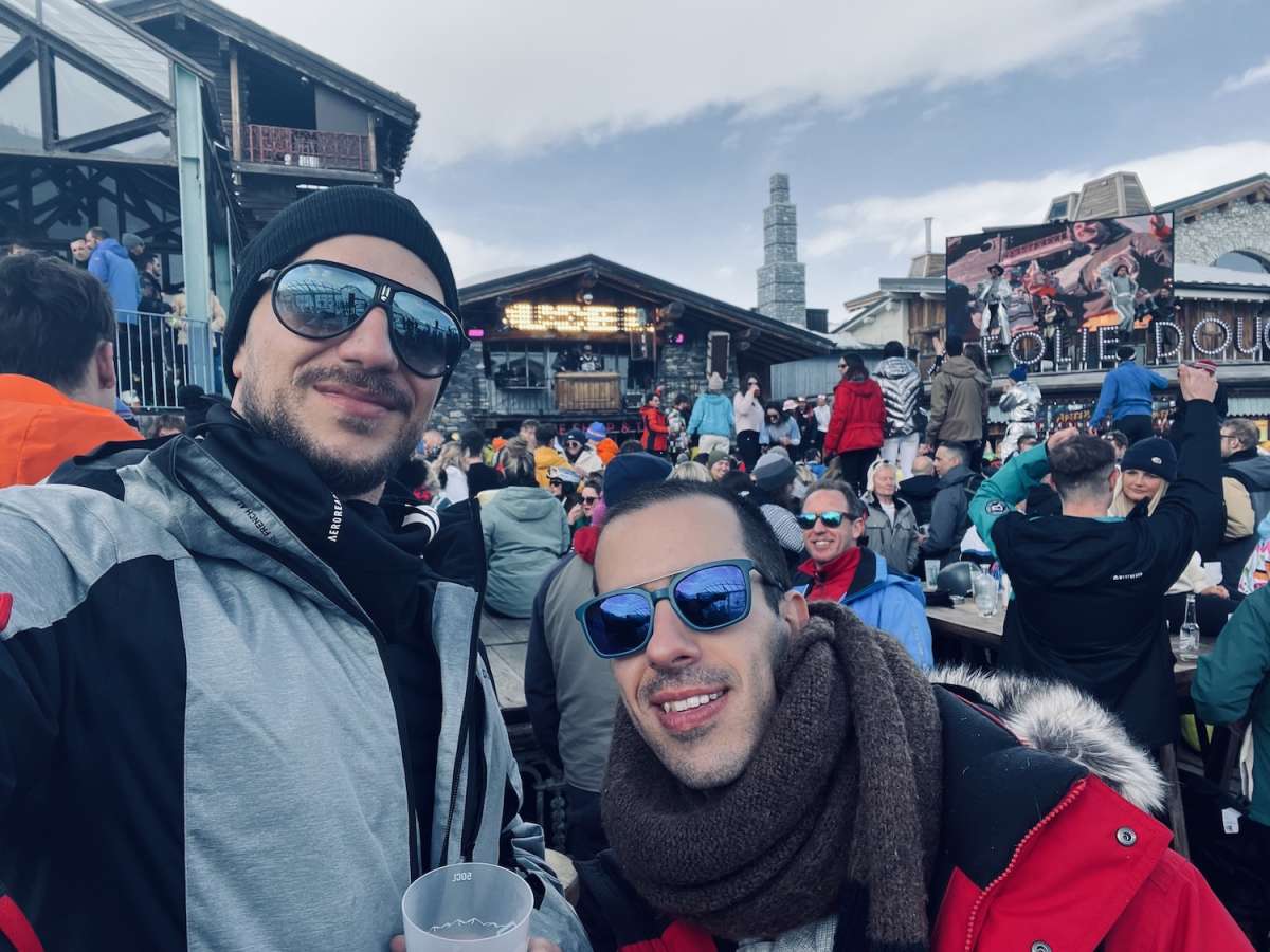 Two men smiling for a photo, basking in the lively atmosphere of European Snow Pride with a bustling Tignes ski resort and mountains in the background.