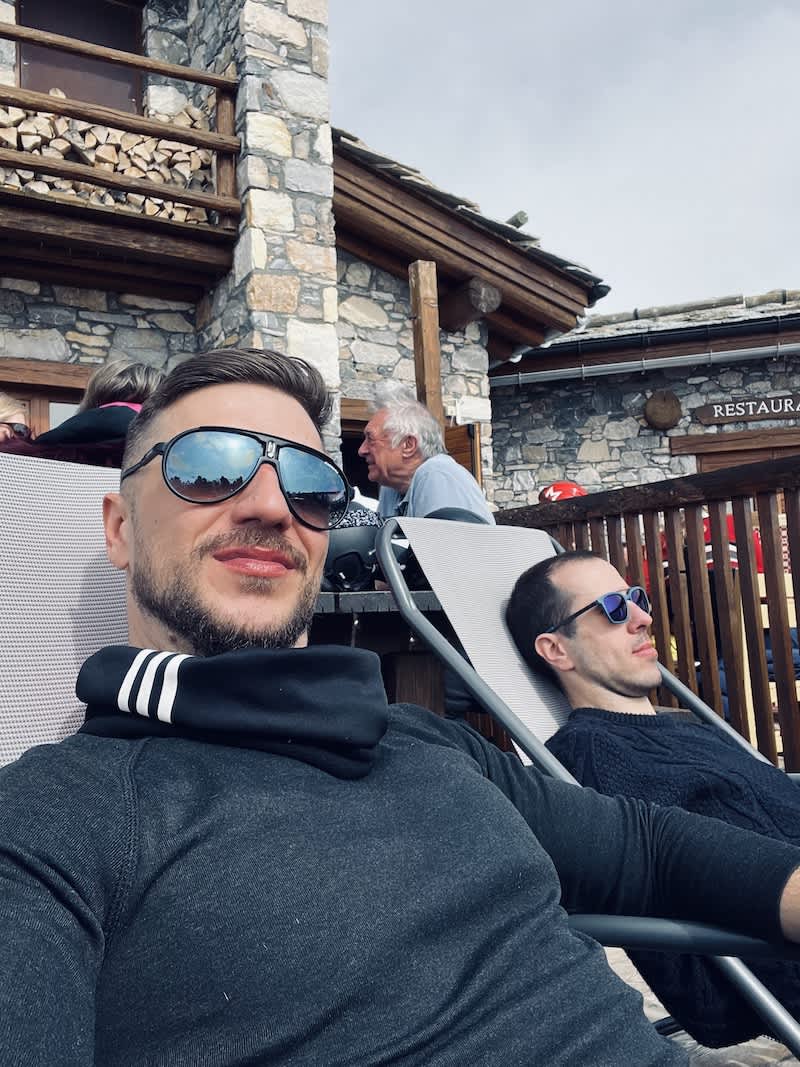 Two men relaxing on lounge chairs, soaking up the sun with content smiles at the European Snow Pride event in Tignes, against a backdrop of picturesque snowy peaks.