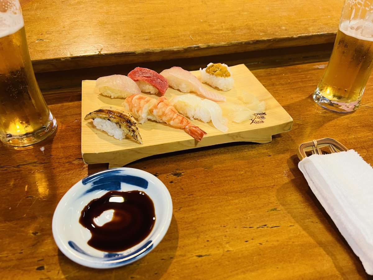 Assorted fresh sushi on a wooden serving board with a side of soy sauce and a pint of beer, ready to be enjoyed in a Nagasaki sushi bar.