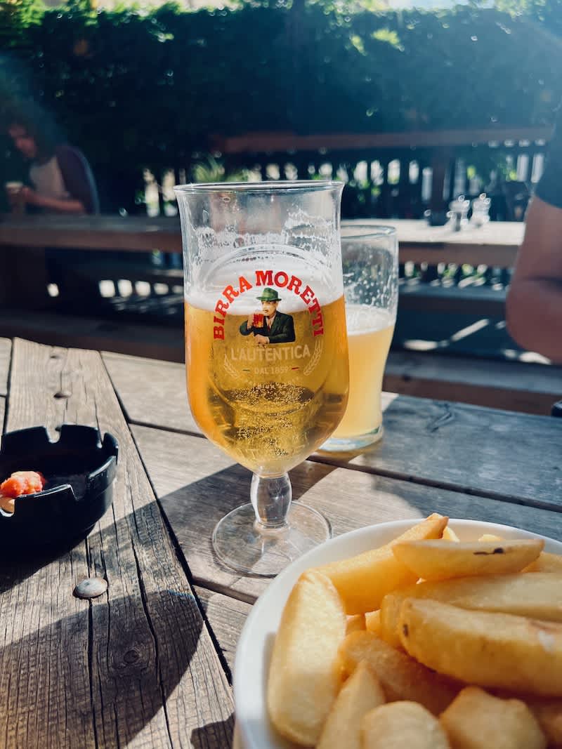 A glass of Birra Moretti beer on a sunny outdoor pub table, accompanied by a bowl of thick-cut chips. The setting is casual with a focus on relaxed dining, representing a typical scene in London's Camden pubs.