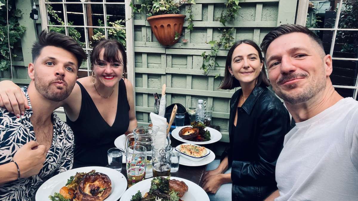 A group of four friends enjoying a meal at The Colonel Fawcett, Camden pub, smiling for a selfie with plates of food visible on the table, exemplifying a friendly, social atmosphere typical of London's vibrant pub scene.