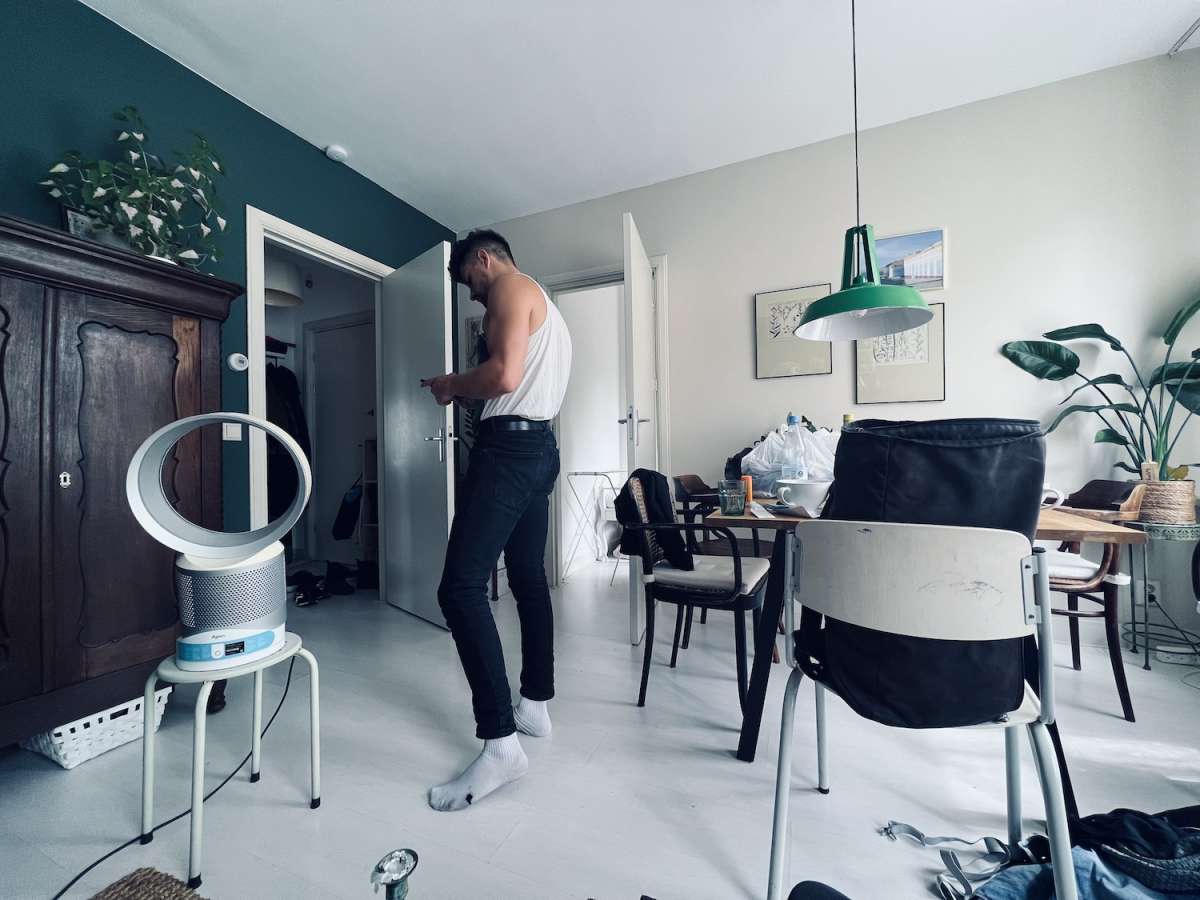 A man standing in a cluttered living room, checking his phone, showcasing a casual and relaxed pre-festival atmosphere typical before heading to events like the Milkshake Festival Amsterdam.