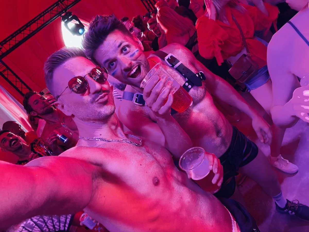 Two smiling men taking a selfie at the Milkshake Festival Amsterdam, surrounded by other attendees and red lighting, highlighting the festive and joyful environment.