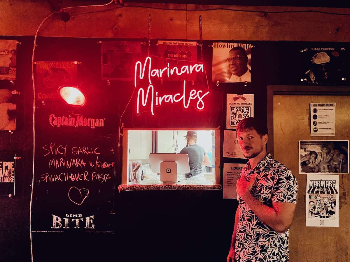 Man giving a thumbs up next to the 'Marinara Miracles' neon sign at a lively Austin, Texas venue, with posters and a menu offering spicy garlic marinara with shrimp, under soft red neon lighting.