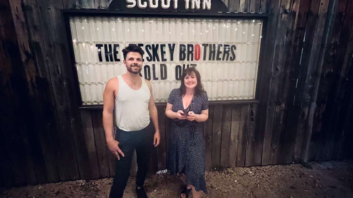 Two friends smiling outside The Historic Scoot Inn in Austin, Texas, under a sign that reads 'The Teskey Brothers,' representing a night out at a live music venue.