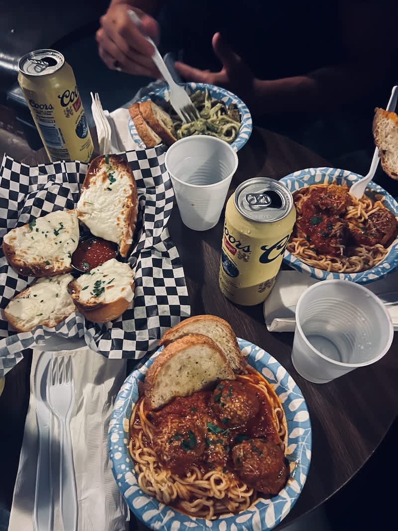 A table filled with various Italian dishes including spaghetti with marinara sauce, garlic bread, and salads, served with cans of beer, highlighting a casual dining scene in Austin.