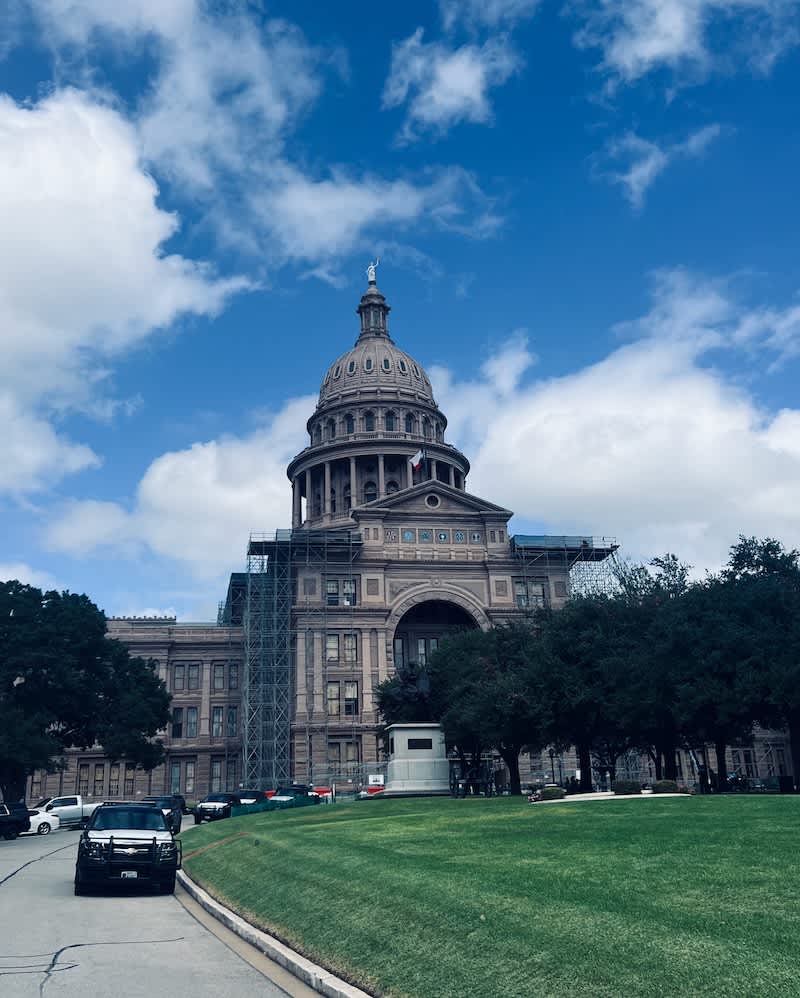 The iconic Texas State Capitol building in Austin under a clear blue sky, surrounded by lush greenery and under renovation with scaffolding visible.