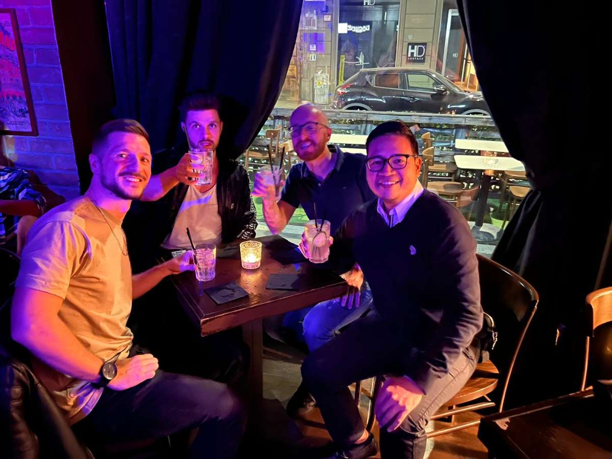 Four smiling men raising their glasses in a toast at a karaoke bar in Milan, enjoying vibrant nightlife with a backdrop of city lights visible through the window.