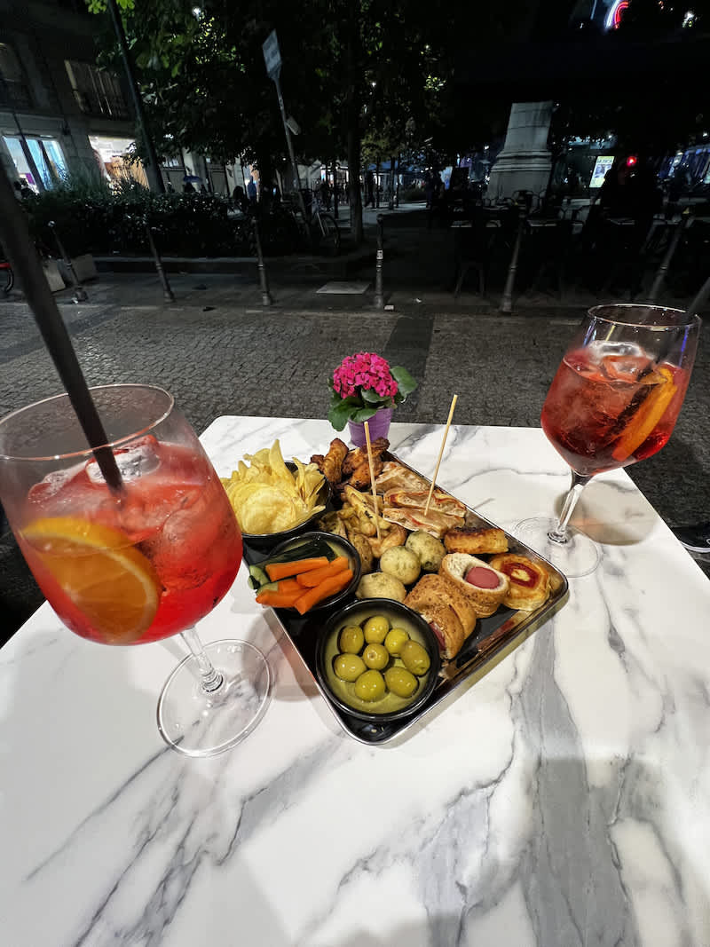 A stylish outdoor table set with an array of aperitivo snacks including olives, chips, and assorted nibbles, accompanied by two large, colorful cocktails, epitomizing Milan's aperitivo culture.