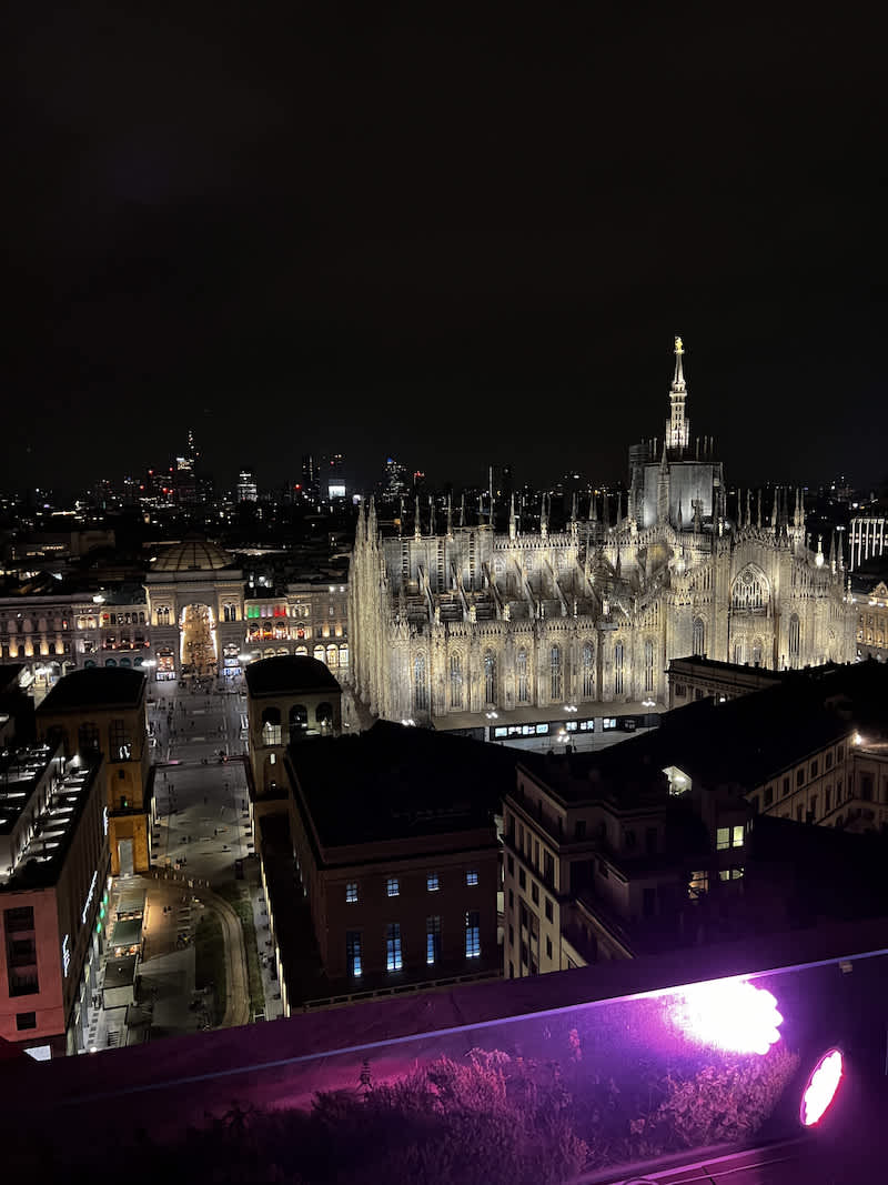 A breathtaking nighttime view of Milan's Duomo, illuminated against the dark sky, with the surrounding cityscape and buildings also lit up, as seen from a high vantage point.