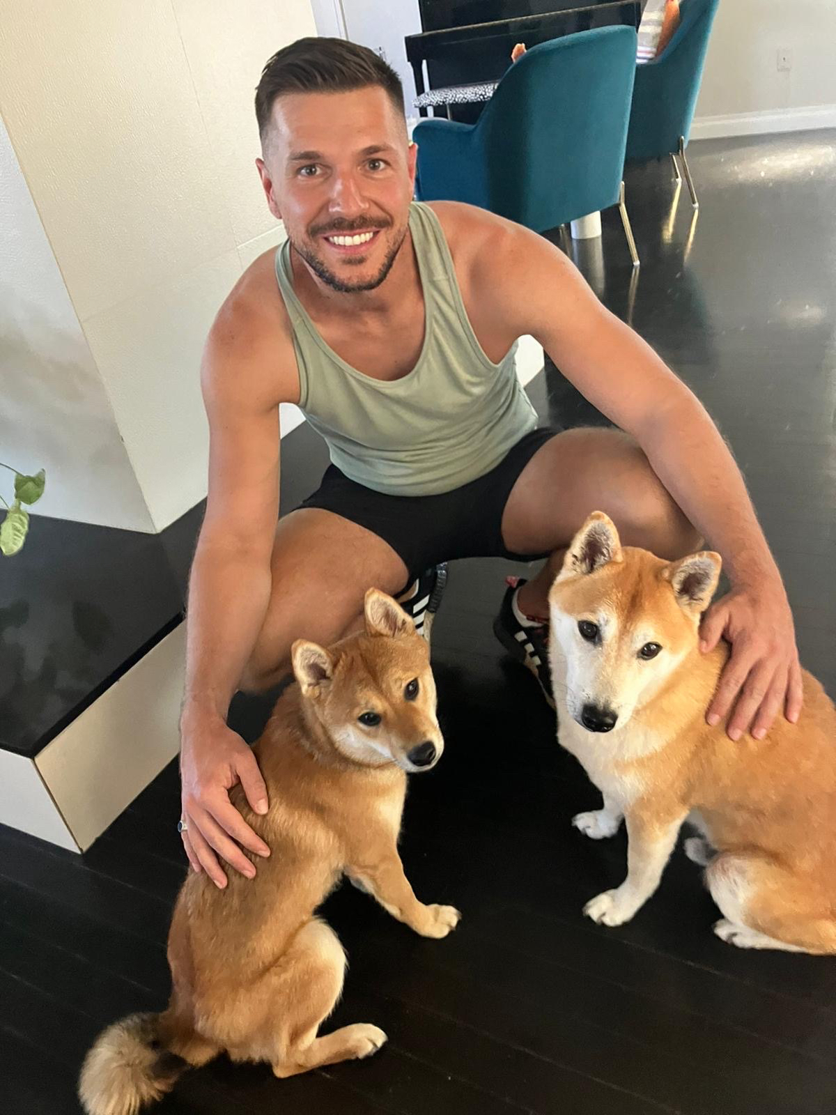 A smiling man in a tank top and shorts is kneeling on the floor with his arms around two Shiba Inu dogs. This image emphasizes the bond between pets and their owner, highlighting the importance of awareness about dangerous foods for dogs.