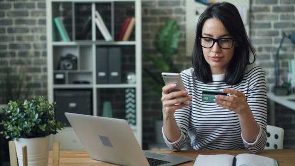 A woman is sat at a table making a payment on her smartphone using a credit card. A laptop is open nearby.