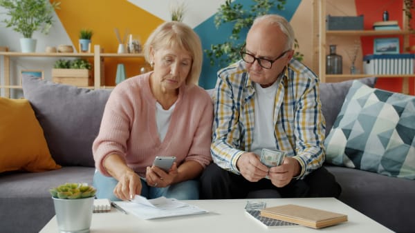 An older couple sat on the sofa looking through bills. The woman is holding a calculator. They both look concerned.