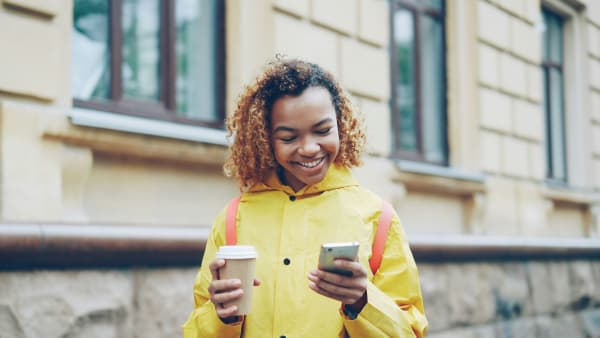 A smiling lady holding her mobile phone in one hand and a takeaway hot drink in the other