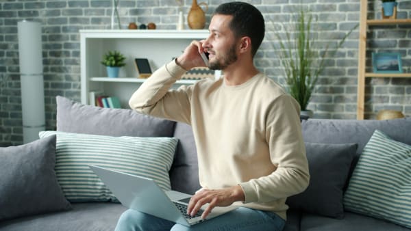 Man sat a grey sofa with a laptop open on his lap. He is talking on a mobile phone.