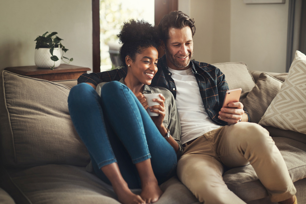 A couple sat on a sofa looking at a smartphone. They are happy and smiling.