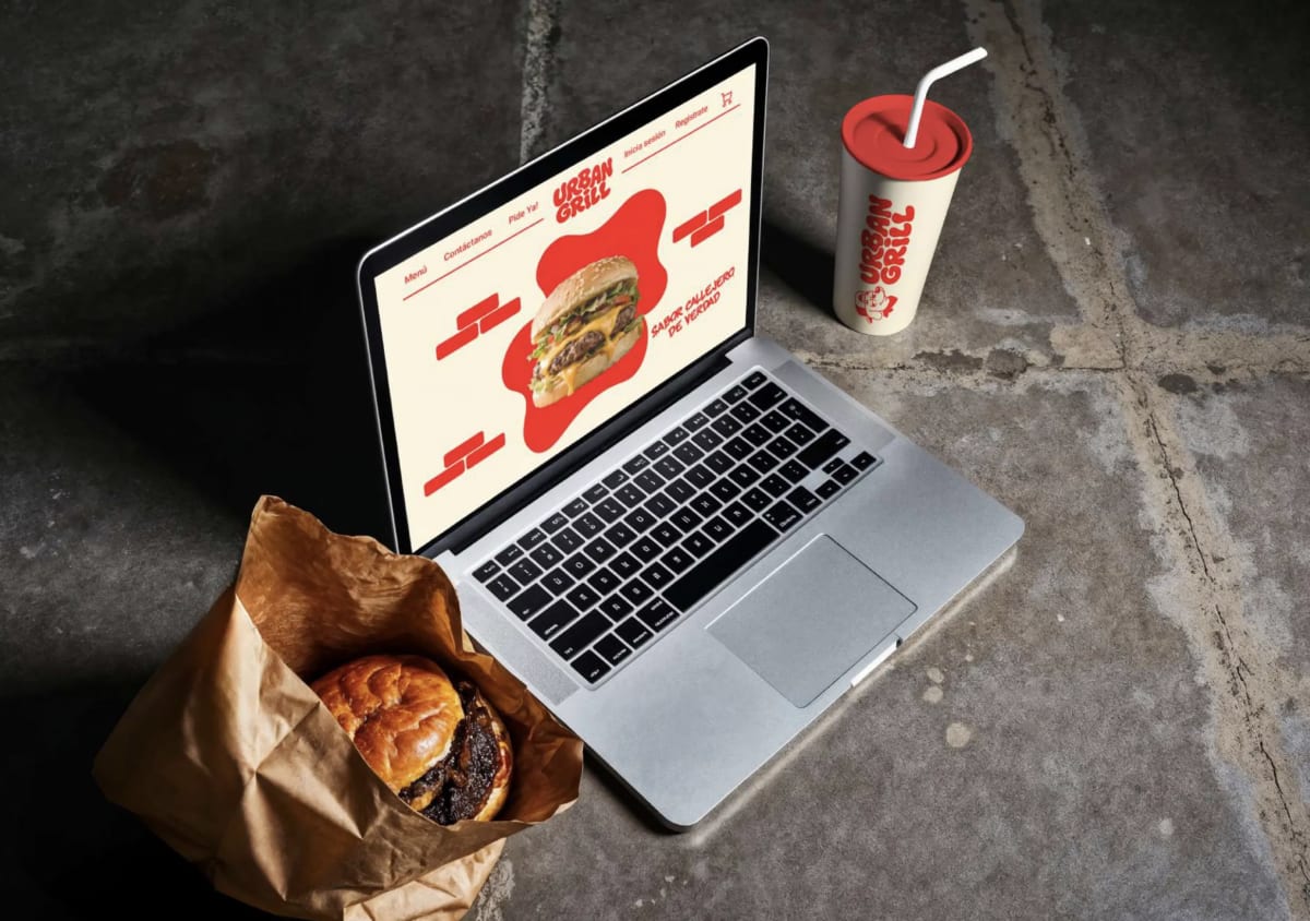 Bodegón cenital de la web de Urban Grill en un portátil con la hamburguesa y el vaso, de Andrea Jurado, alumna de Diseño Gráfico y Motion Graphics en TRAZOS