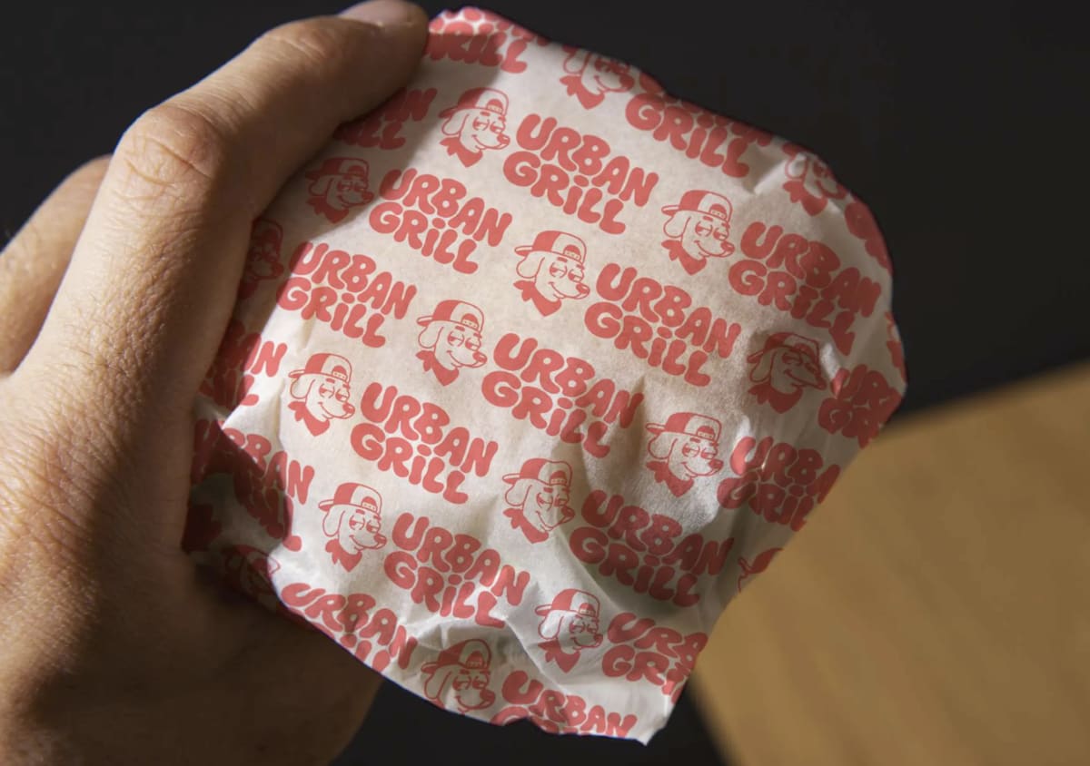 Hamburguesa envuelta en el papel estampado de Urban Grill, sostenida con la mano, de Andrea Jurado, alumna de Diseño Gráfico y Motion Graphics en TRAZOS