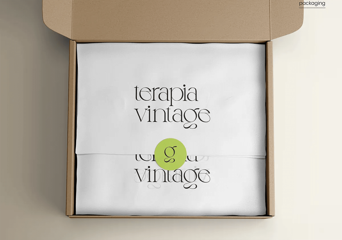 Caja de envío de cartón abierta con prendas dobladas y pegatina de marca de Terapia Vintage, de Aida Domínguez, alumna de Diseño Gráfico y Motion Graphics en TRAZOS