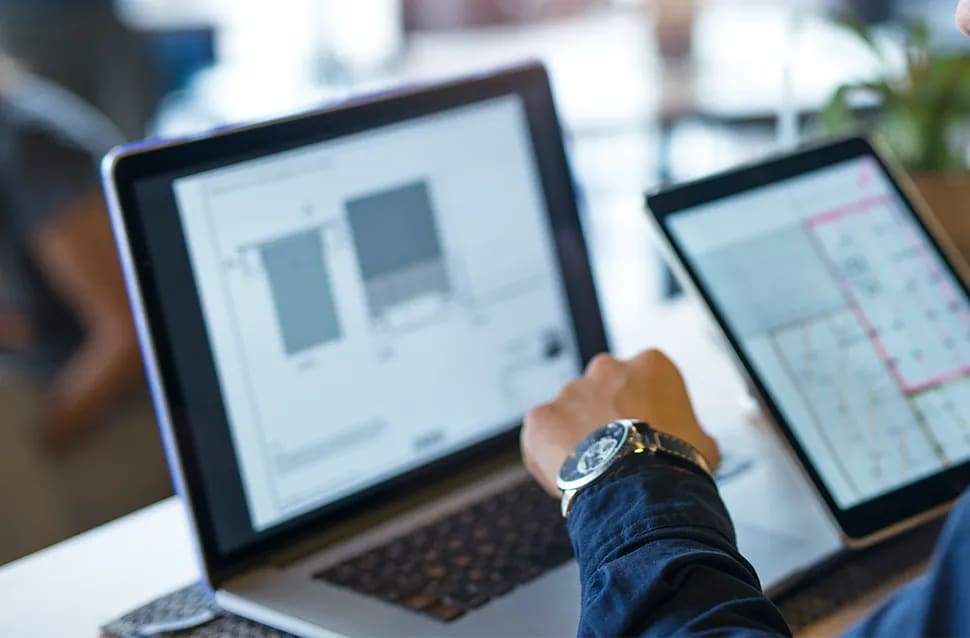 Persona trabajando en un portátil con diseños de interfaz junto a una tablet con una cuadrícula de proyecto, entornos reales y automatización, módulo del Máster en Desarrollo Web Full Stack con React, Node.js e IA de TRAZOS, escuela de arte digital en Madrid