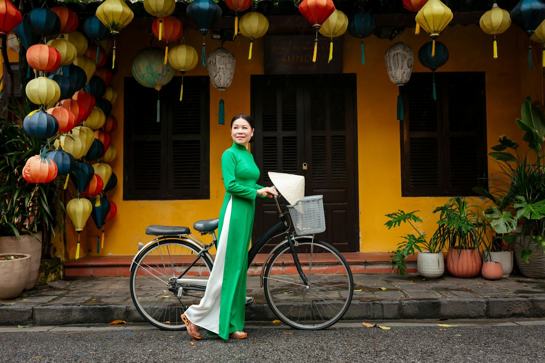 A woman in a green ao dai stands with a bicycle in colorful Hội An.
