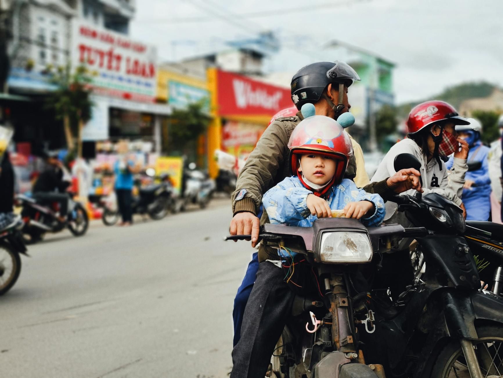 Young child on motorcycle with adult in vibrant street setting, helmets on.