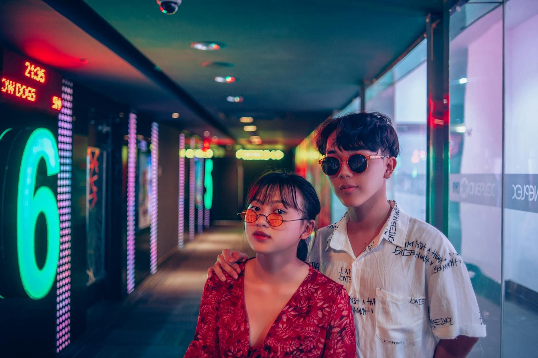 Young couple in sunglasses stands stylishly in a neon-lit movie theater hallway.