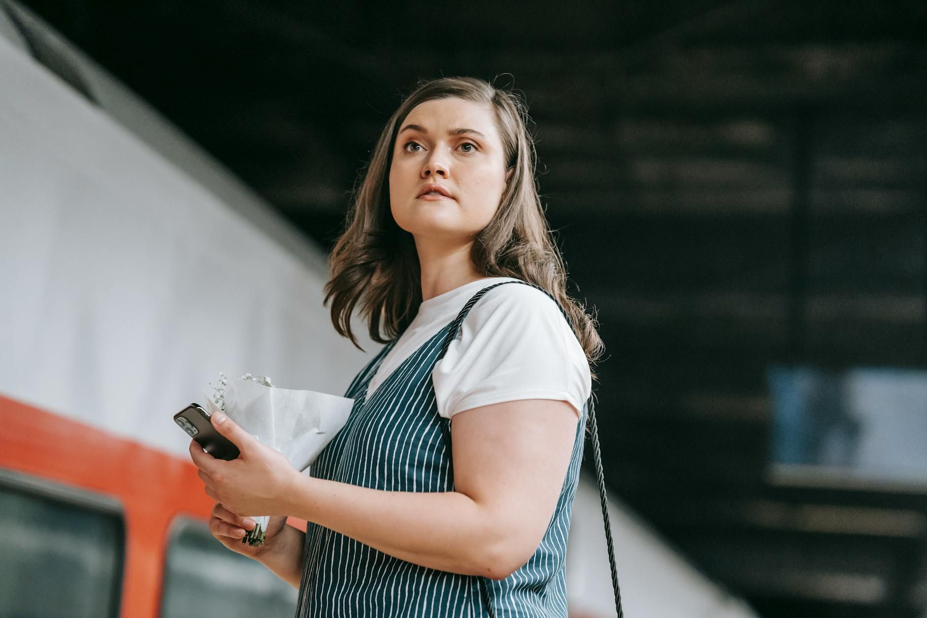 Young woman using phone at a train station, waiting for departure under dim lighting.