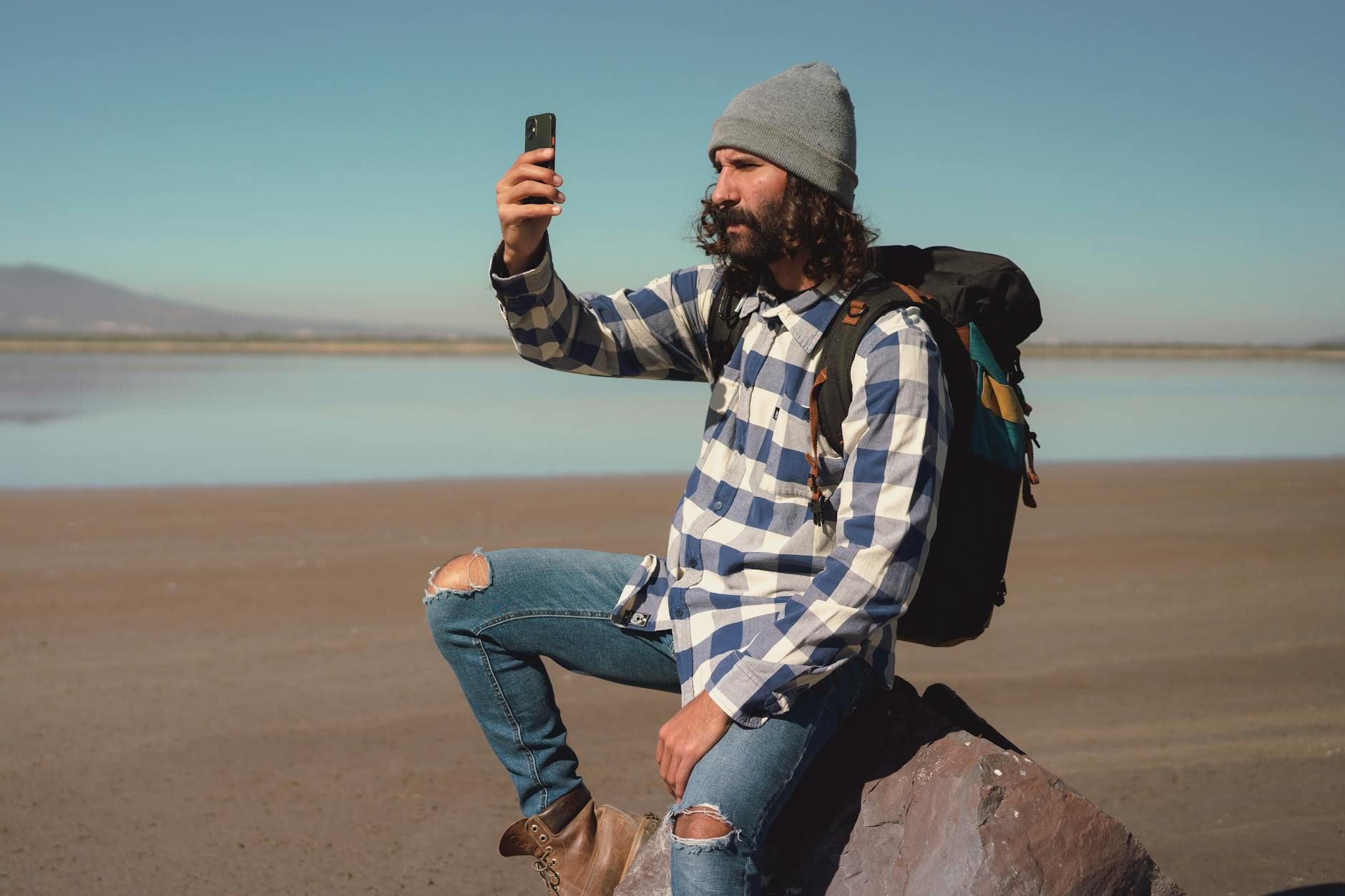 Casual man in trendy outfit takes selfie during outdoor adventure by a lake.